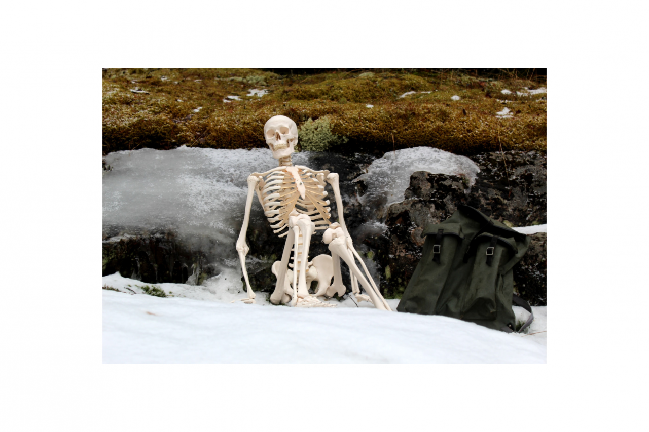 Human skeleton sitting on snow-covered ground next to their backpack enjoying the view on their break from a stroll outdoors