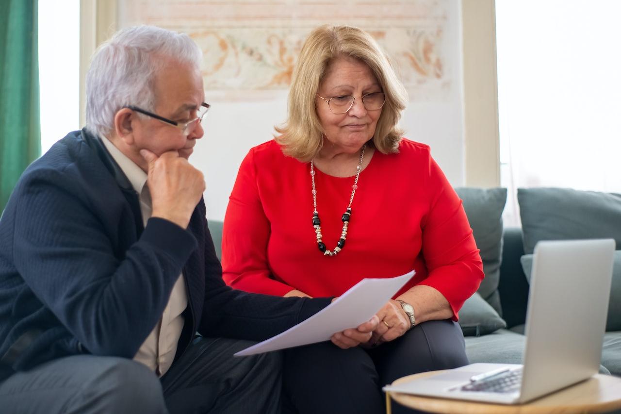 Older man and older woman looking at paperwork and their laptop.