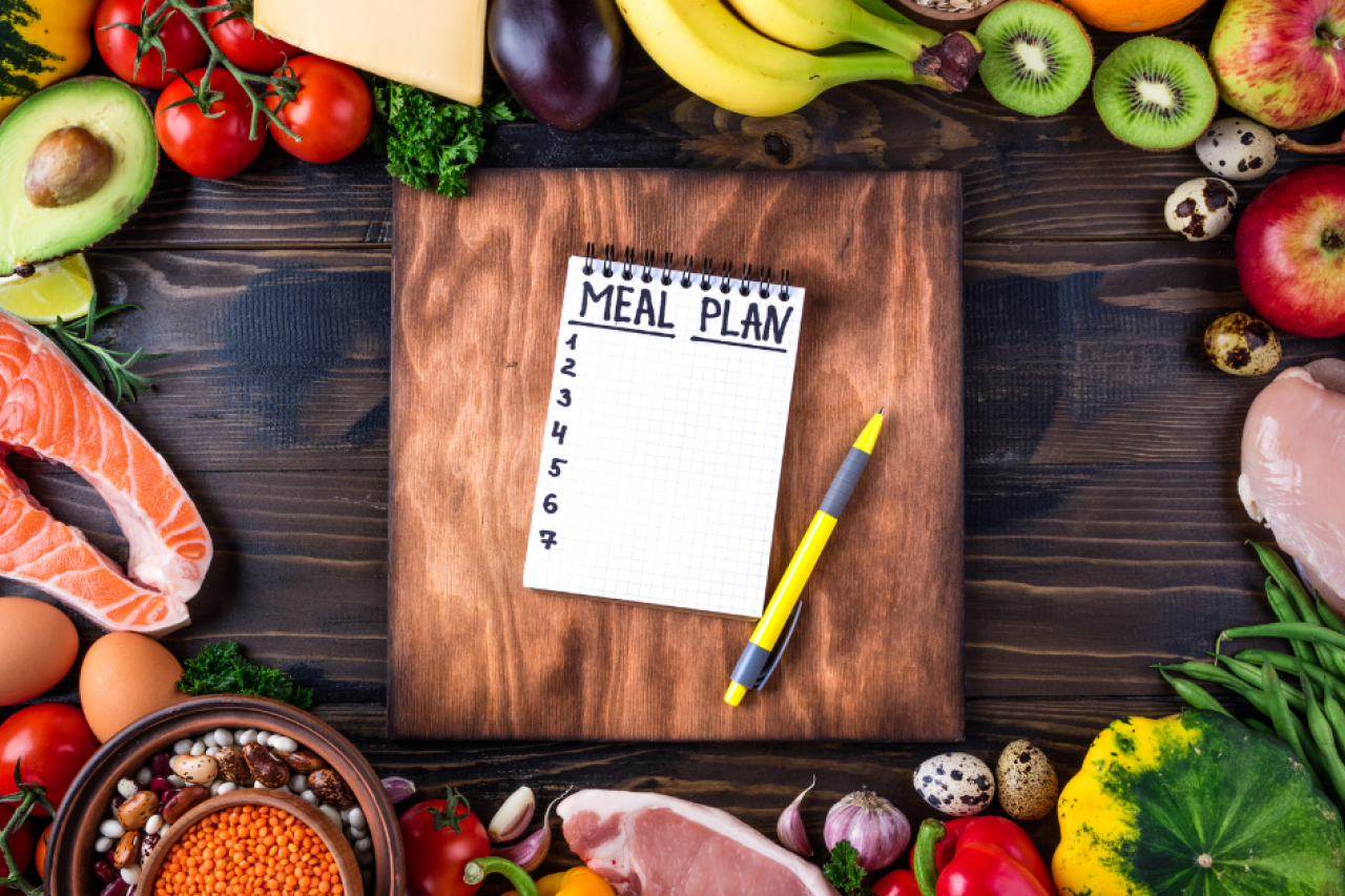 A wooden table layout with healthy foods such as fresh fruits and vegetables, different kinds of meat, nuts, and grains along the edges, and a notebook with a “meal plan” list written on it next to a pen in the center on top of a wooden cutting board