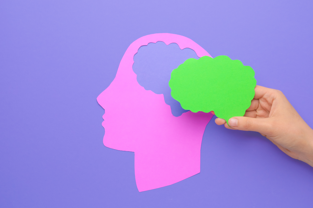 A hand holding a paper cutout of a human brain over the paper cutout of a human head with a pastel purple color background