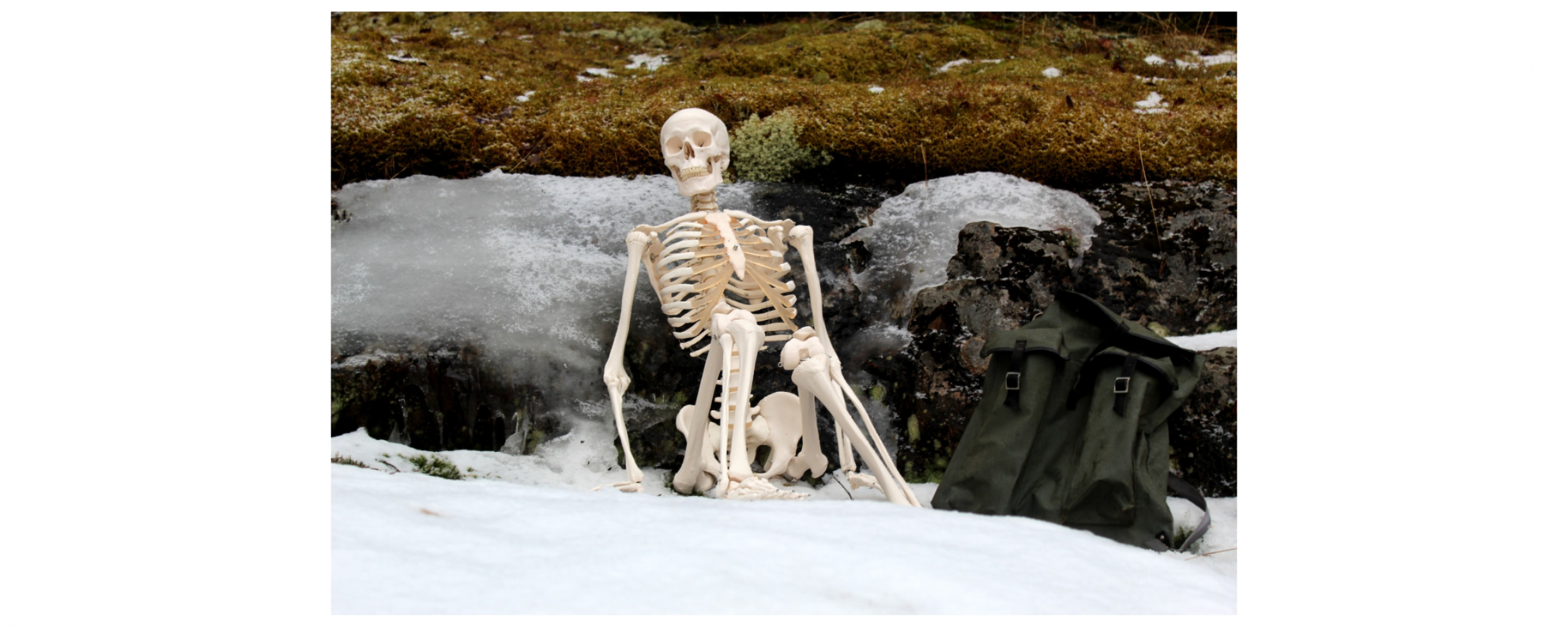 Human skeleton sitting on snow-covered ground next to their backpack enjoying the view on their break from a stroll outdoors