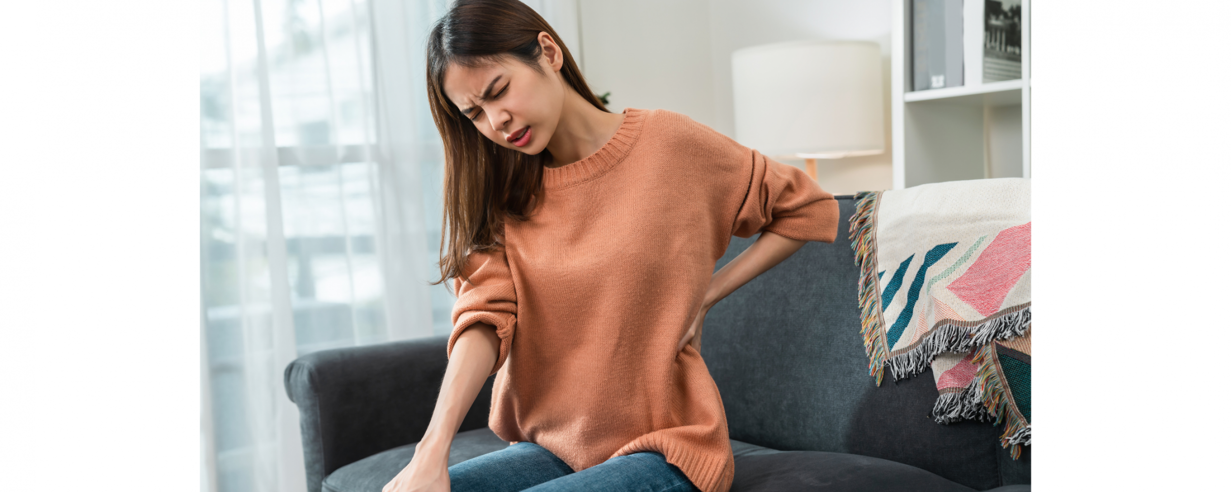 Woman sitting on couch with hand on waist/lower back wincing in pain