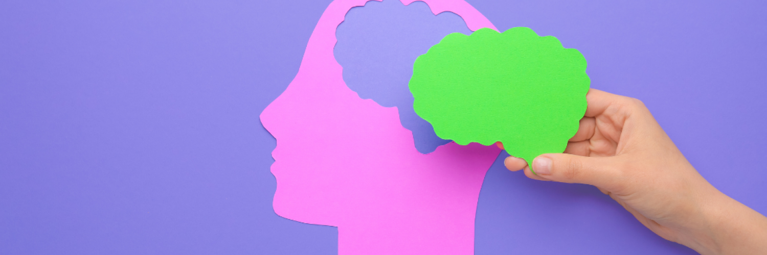 A hand holding a paper cutout of a human brain over the paper cutout of a human head with a pastel purple color background